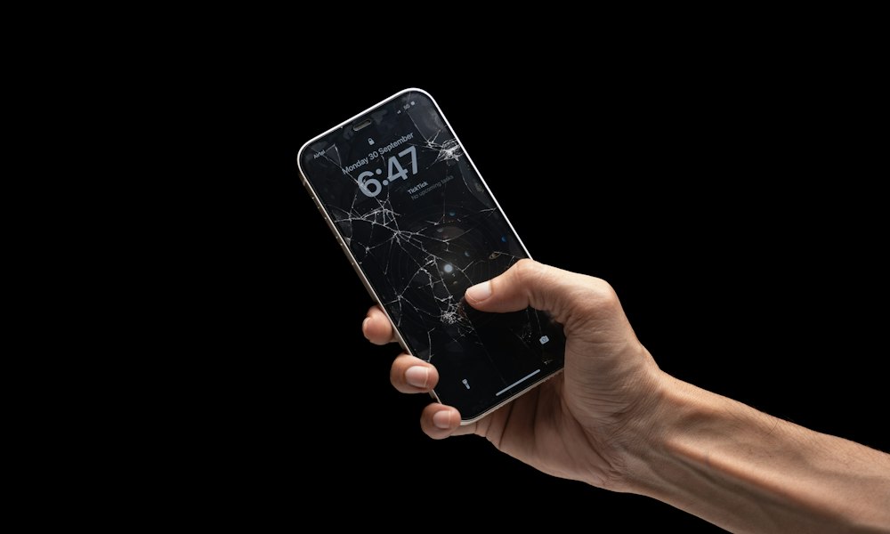 Mano con un celular de pantalla trizada sobre fondo negro en su comuna